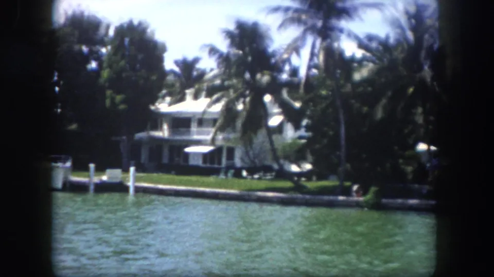 8mm home-movie frame circa 1960, Tampa, Florida — two-story white waterfront home with balcony, canal dock pilings, and tropical palms; small figure appears near the waterline.