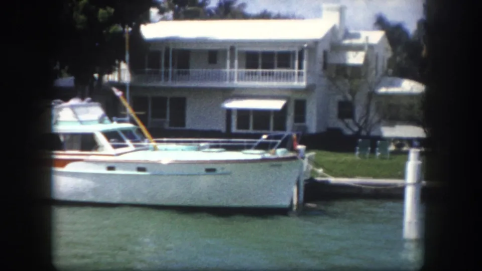 Classic 1960s-era white cabin cruiser moored at a private dock in Tampa, Florida — two-story waterfront home with balcony and tropical palms behind; figure near stern area.