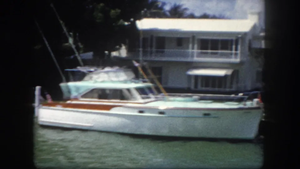 Early 1960s cabin cruiser with teal cabin and wood-trim hull on a Tampa-area canal; two-story white waterfront house, balcony, and sailboat mast visible behind, 8mm film
