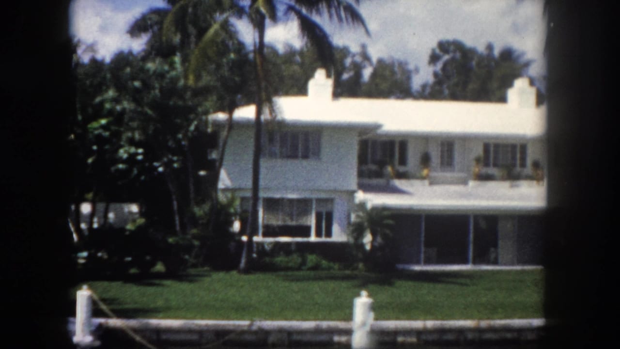Mid-century white waterfront residence with large windows, dock piling, and manicured canal-side lawn in Tampa, Florida, c.1960; palm trees in background, 8mm film