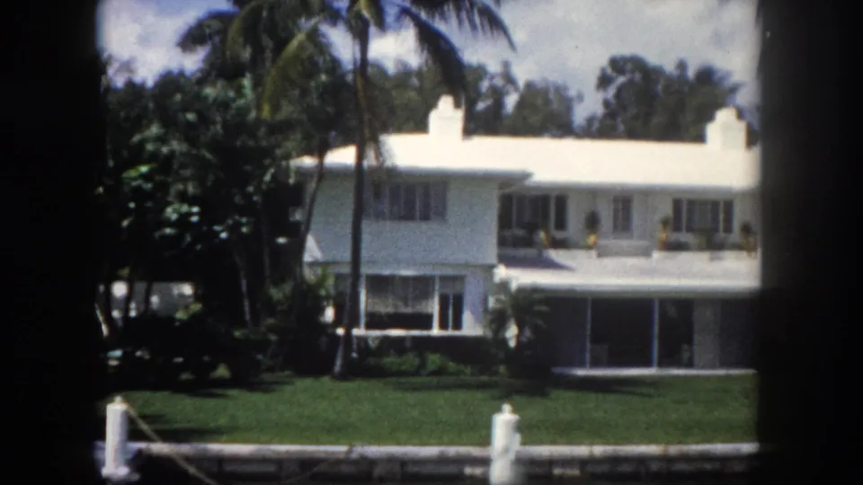 Mid-century white waterfront residence with large windows, dock piling, and manicured canal-side lawn in Tampa, Florida, c.1960; palm trees in background, 8mm film