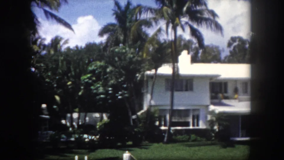 Tall palm trees and dense tropical landscaping before a white mid-century Florida waterfront home, Tampa area, c.1960; partly cloudy sky, 8mm film grain visible