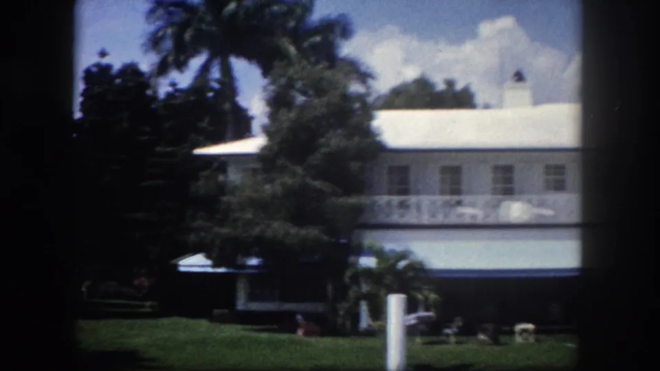 8mm film frame showing a wide-angle view of a white Florida residence receding behind palm trees and tropical landscaping, circa 1960 Tampa.