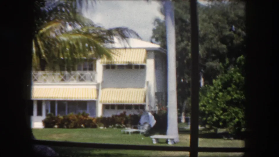 8mm home movie, c.1960 Tampa, Florida — frontal view of a mid-century white residential building with striped awnings on both floors, set in a tropical garden.