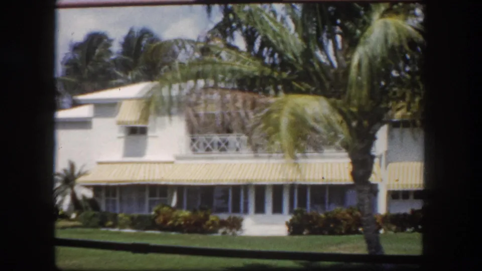 White stucco Florida house with yellow-and-white striped awnings and palm trees, seen through a window, Tampa, c. 1960.