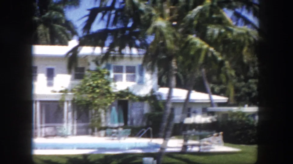 Residential or resort swimming pool area with a white building and palm trees, seen through a window in 1960s Florida.