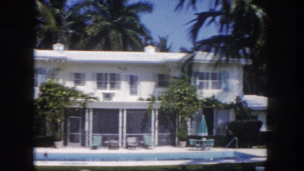 White two-story Florida building with screened lanai panels, rectangular swimming pool, patio umbrella and chairs, palm trees, circa 1960