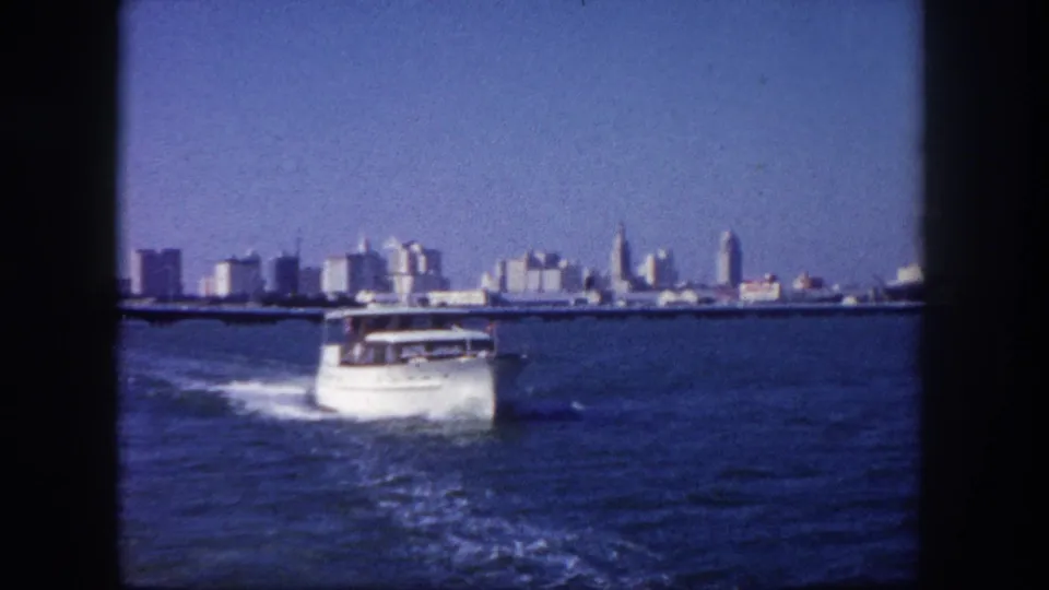 White cabin cruiser motorboat on open bay water leaving a wake, with a mid-century urban skyline on the horizon, possibly Tampa, circa 1960