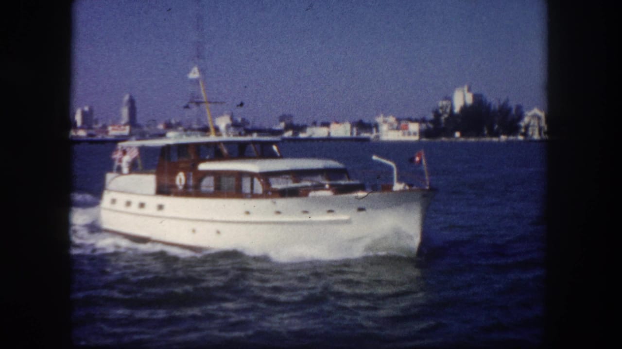White motor yacht with wood-trim cabin cruising through coastal waters, low shoreline with trees and buildings visible behind.
