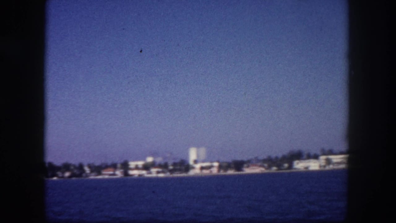 Boat-level view of a coastal shoreline with low- and mid-rise white buildings, likely Tampa Bay area, circa 1960, 8mm color film.