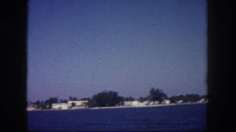 Open water view with a low-lying tree-lined shoreline and scattered white structures, likely Tampa Bay area, circa 1960, 8mm color film.