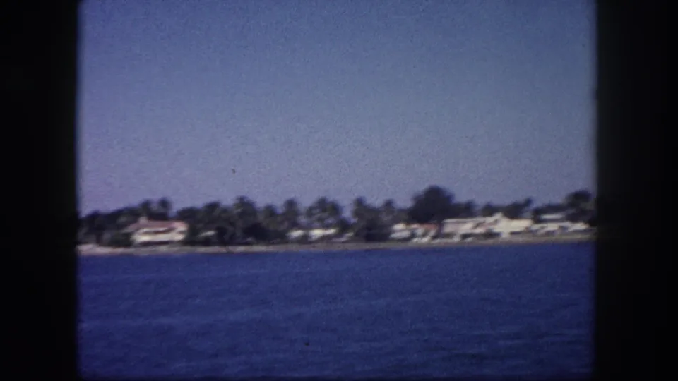 8mm home movie frame of a Florida coastal community seen from a moving boat, with palm trees and low-rise waterfront buildings, circa 1960.