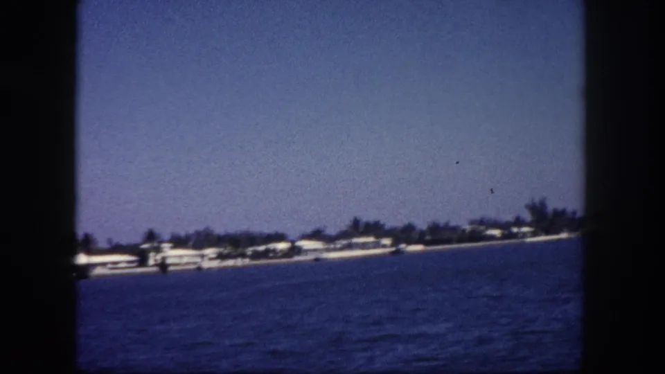 8mm frame showing a widening view of a Florida coastal strip from a moving boat, palms and low buildings receding across open water, circa 1960.