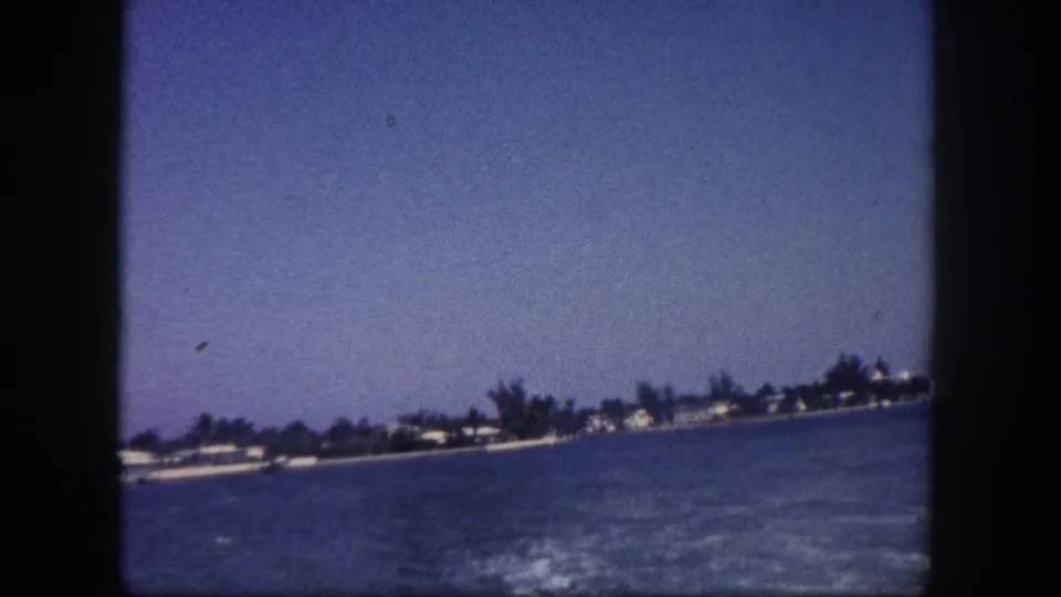 8mm home movie frame of a receding Florida waterfront seen from a boat, with palm trees and low-rise structures at the far shore, circa 1960.