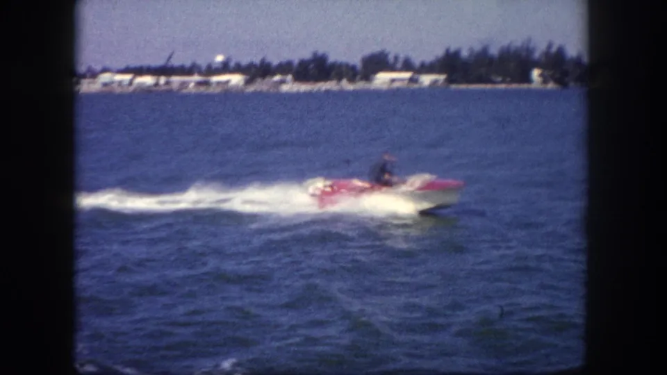 Red speedboat with visible operator crossing open water near Tampa Bay, circa 1960, filmed on 8mm; wooded shoreline and low buildings in background.
