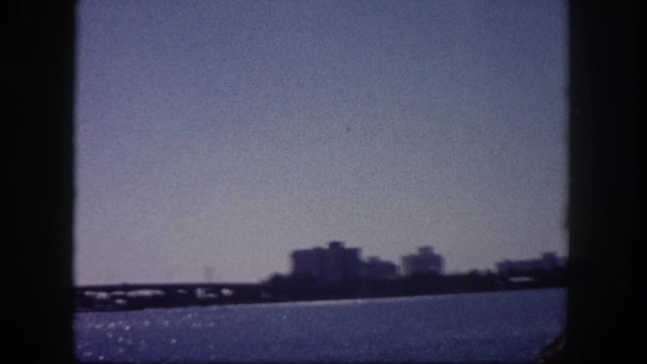 1960 8mm home movie frame showing Tampa Bay waterfront, distant low-rise skyline, and a bridge structure under a hazy sky.