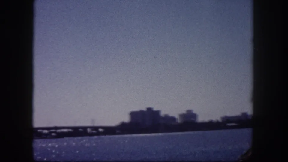 1960 8mm home movie frame showing Tampa Bay waterfront, distant low-rise skyline, and a bridge structure under a hazy sky.