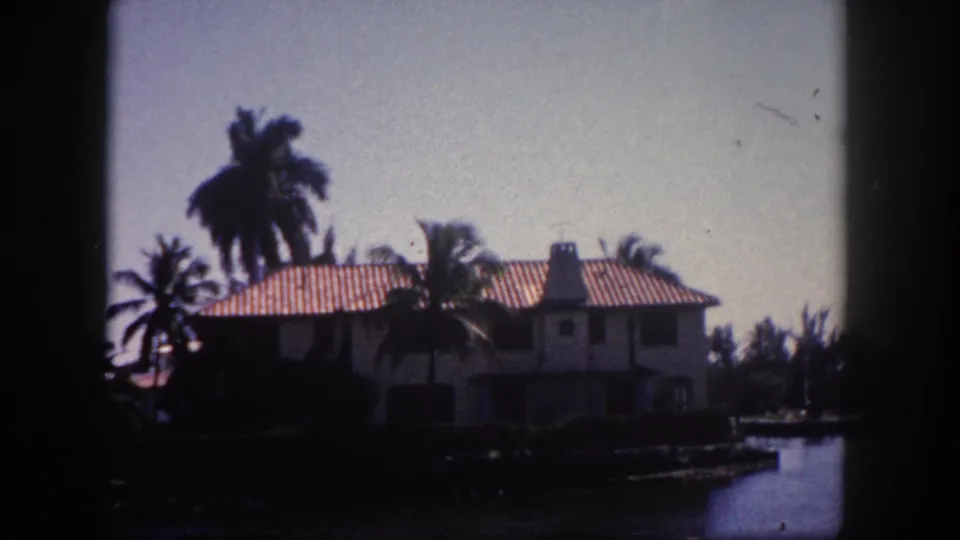 1960 8mm frame showing a stucco residence with red terracotta roof tiles and tall palm trees on the Florida waterfront.
