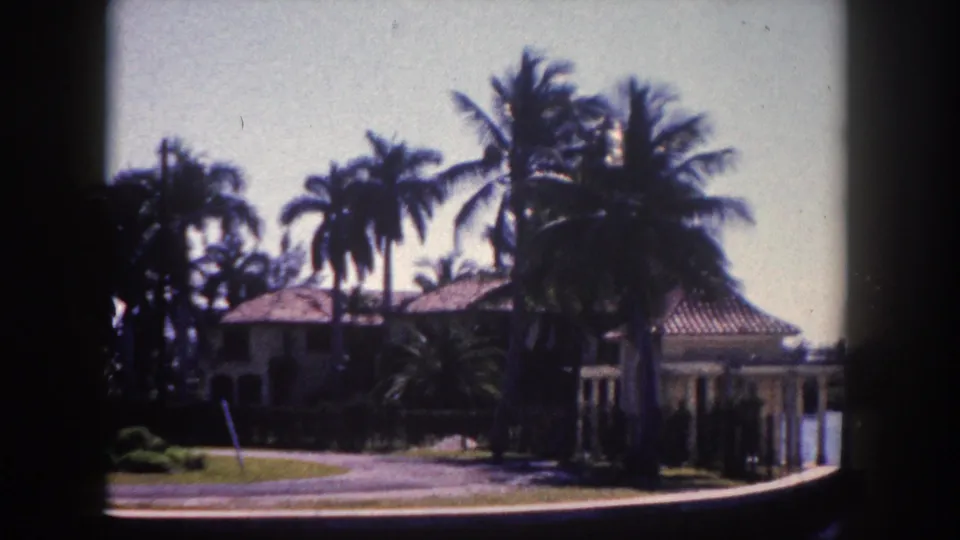 Wide shot showing terracotta-roofed house and columned gazebo among dense palm trees with a road in the foreground; 8mm footage, Tampa, Florida, circa 1960.