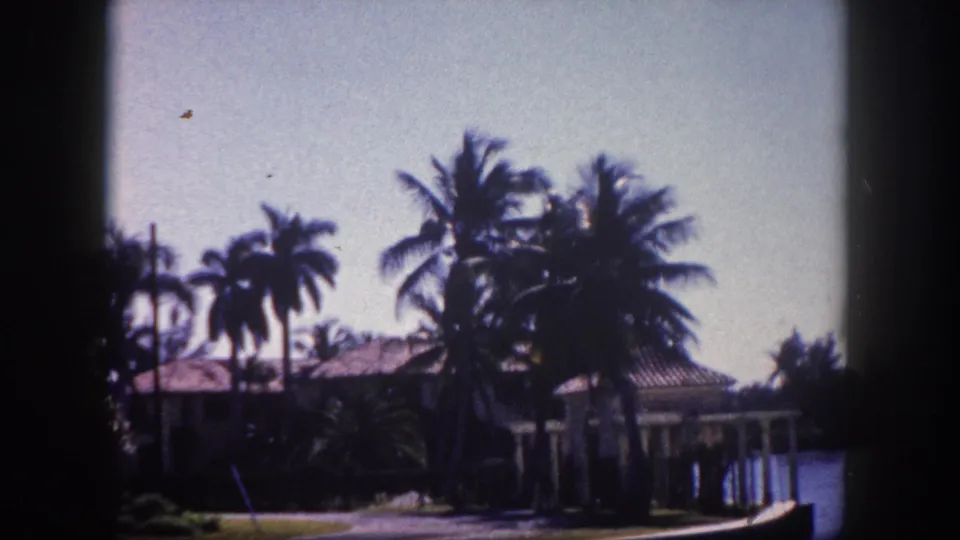 8mm home movie frame of a waterfront property with a tile-roofed building and tall palms, likely a Tampa Bay area waterway, circa 1960.