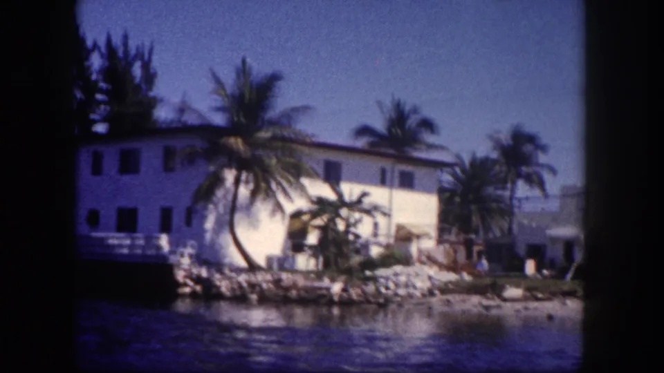 8mm home movie frame of a white waterfront building with palm trees and rocky seawall, likely a Tampa Bay area waterway, circa 1960.