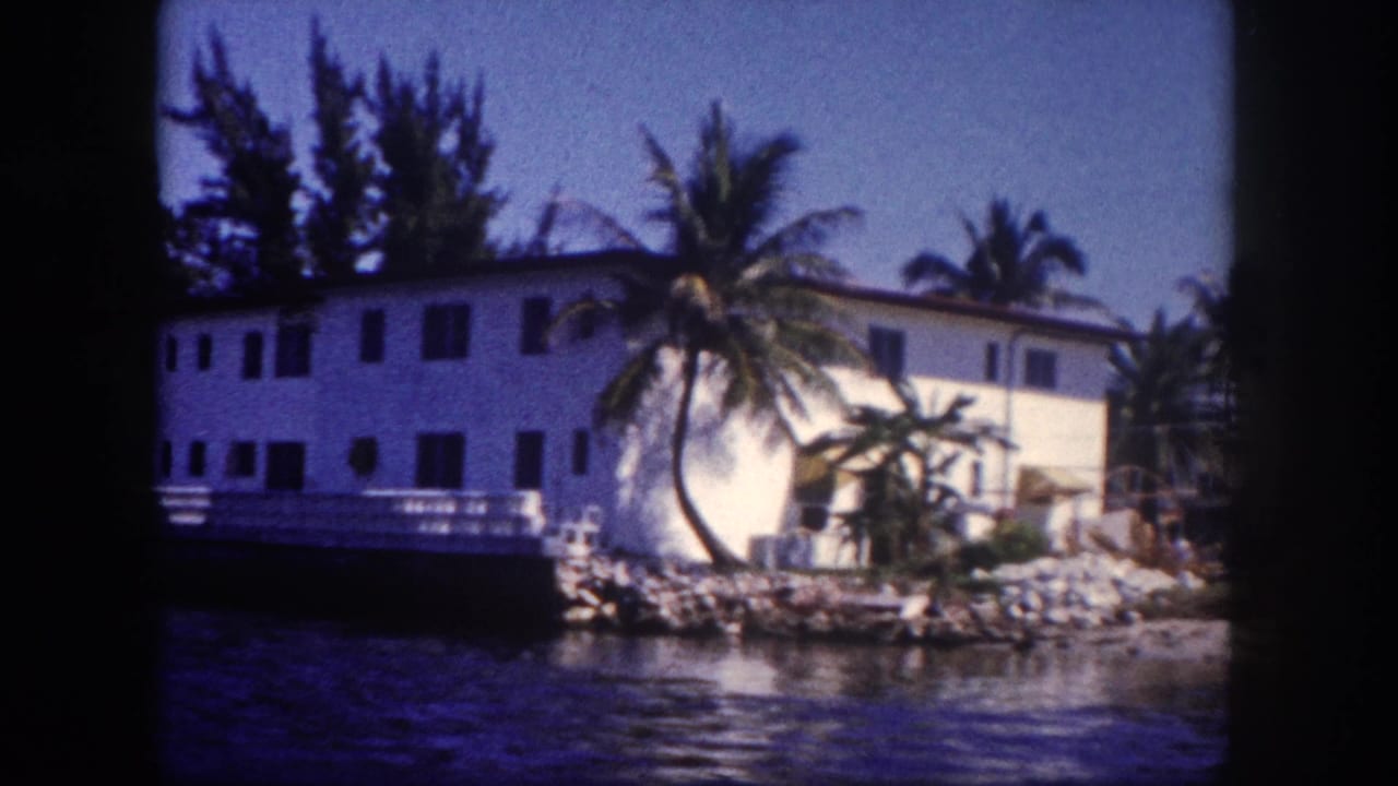 8mm home movie frame, c. 1960, Tampa, Florida: white waterfront building with railed porch, palm trees, rocky seawall, and blue waterway in foreground.