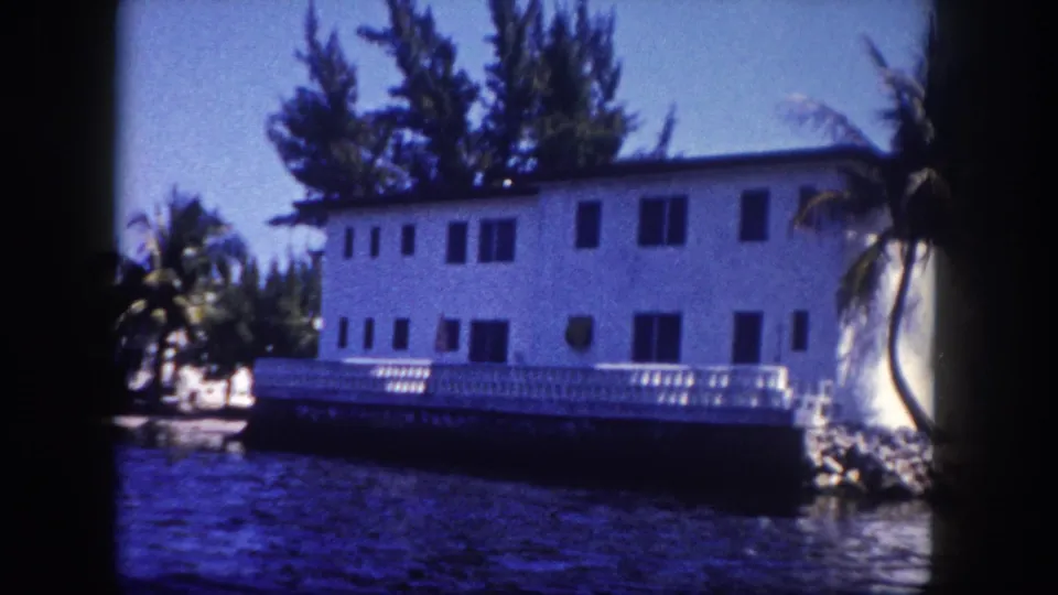 8mm frame, c. 1960, Tampa, Florida: two-storey white building with white porch railing, tall pines at left, palm trees, calm waterway in foreground.