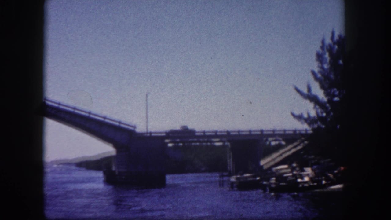 Bascule drawbridge spans raised over a waterway in Tampa, Florida, circa 1960; structural or dock elements visible at right edge, tree line at upper right, 8mm film.