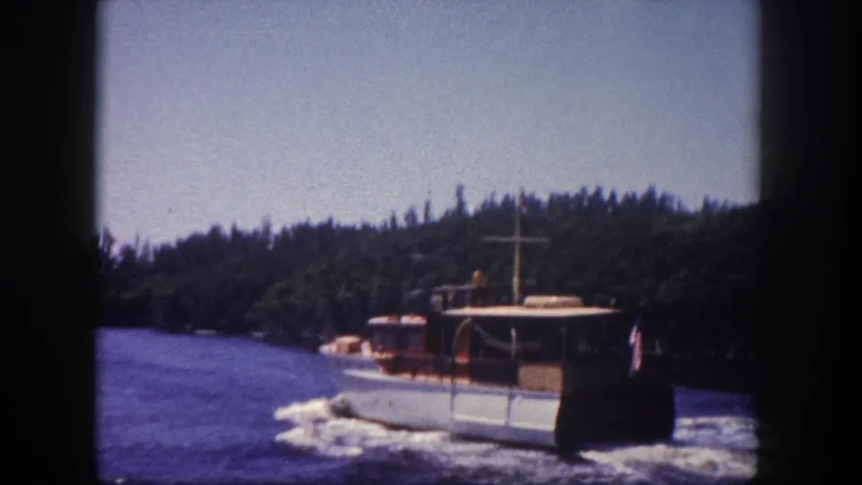 Mid-century white-hulled cabin cruiser with wooden trim moving through a Florida waterway, tree-lined shore in background, apparent stern flag, shot on 8mm film circa 1960 in Tampa.