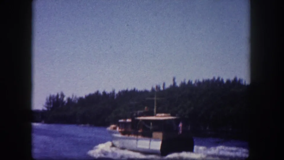 White-hulled cabin cruiser continuing along a Florida waterway flanked by dense tree-lined shore, shot on 8mm film circa 1960 in Tampa.