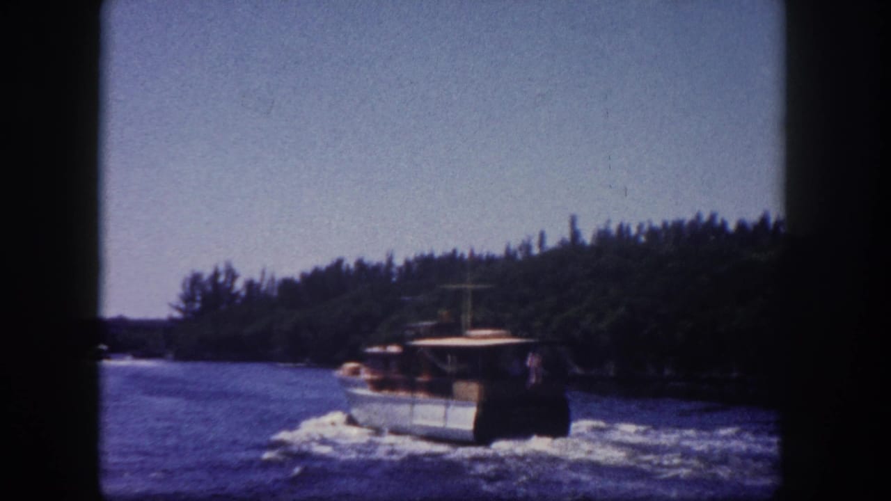 A brown-hulled harbor workboat moving through blue water with a dense tree-lined shore behind it.