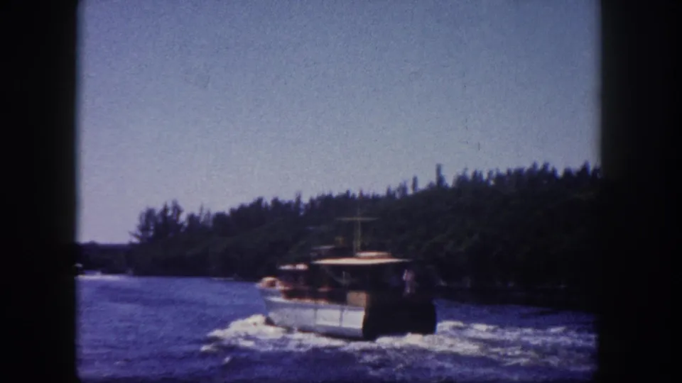 Small harbor workboat with cabin moving through a Florida coastal waterway, dense tropical treeline visible; 8mm color film, circa 1960.