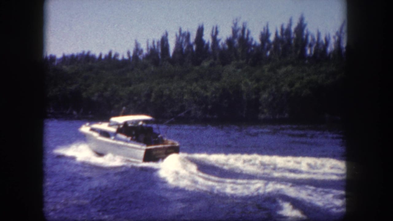 White hardtop cabin cruiser underway at speed on a Florida coastal waterway, large foaming wake, tropical treeline and blue sky; 8mm color film, circa 1960.