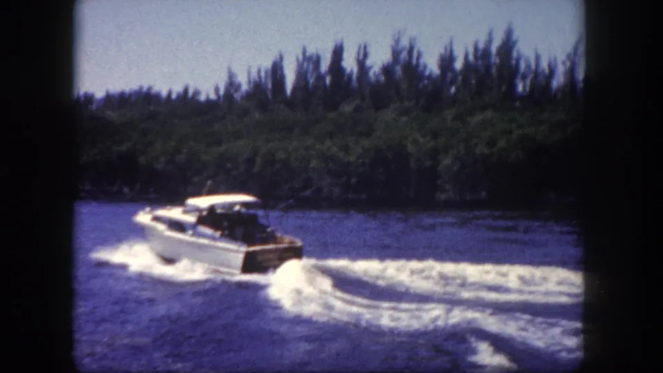 White hardtop cabin cruiser underway at speed on a Florida coastal waterway, large foaming wake, tropical treeline and blue sky; 8mm color film, circa 1960.