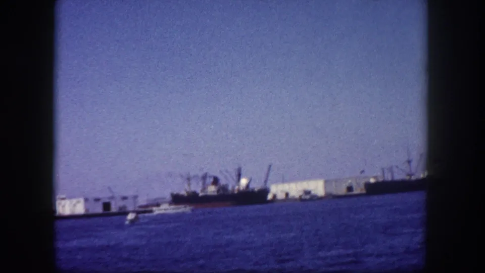 Wide view of a harbor with cargo vessels, industrial warehouses, and port cranes, possibly Tampa Bay area; 8mm film, circa 1960.