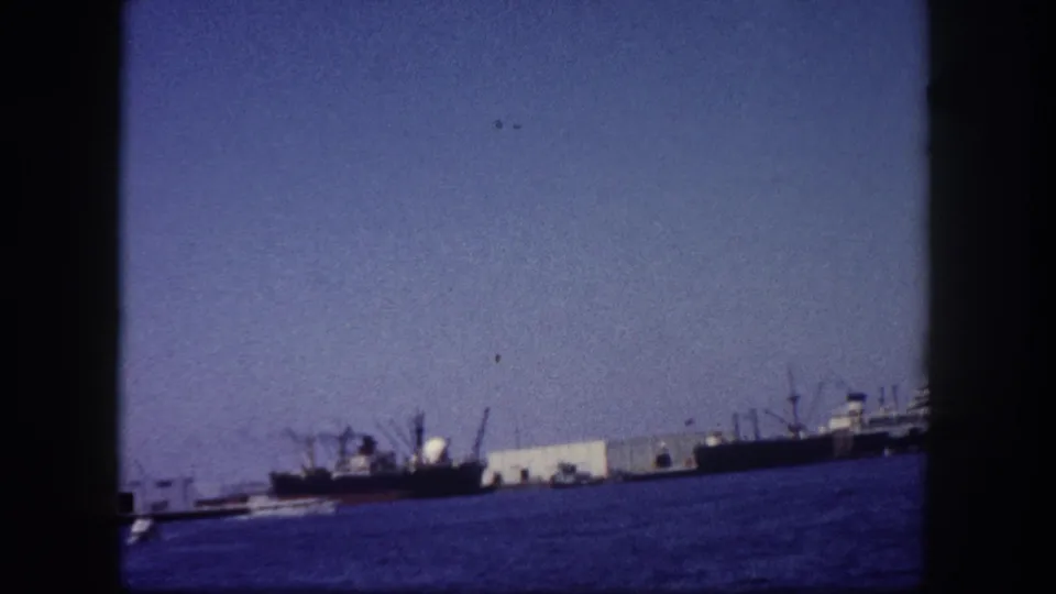 Continued wide harbor shot with cargo freighters, port cranes, and waterfront industrial buildings, possibly Tampa Bay area; 8mm film, circa 1960.