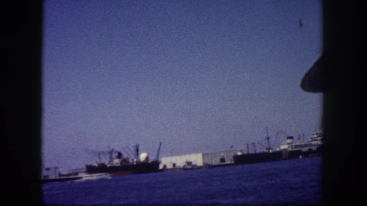 Cargo ships docked at a waterfront pier, shot from water level on 8mm film, circa 1960.