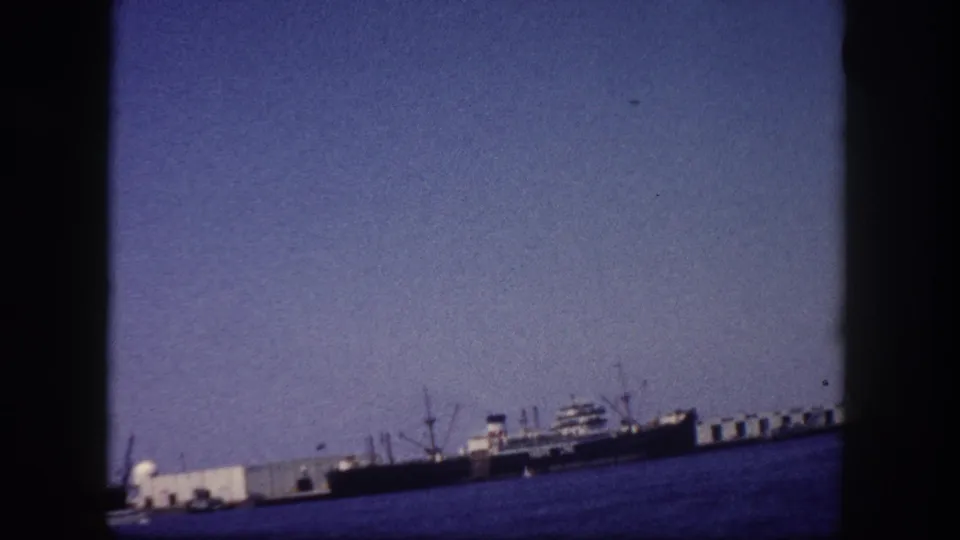 Tampa, Florida harbor circa 1960, cargo vessels and pier infrastructure visible along the waterline under a clear sky on 8mm home-movie film.