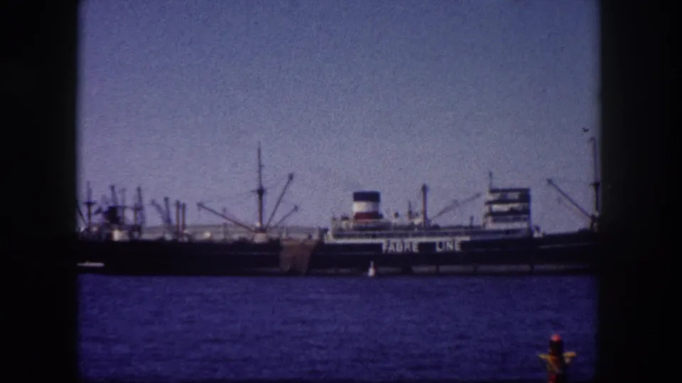 Fabre Line passenger-cargo liner docked at Tampa port, Florida, circa 1960; orange buoy in foreground, loading cranes visible, 8mm film grain and blue cast.