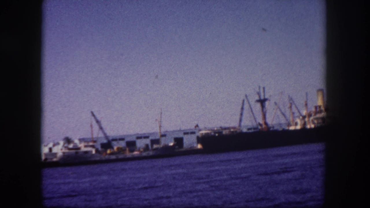Cargo ship moored at Tampa port with loading derrick booms, circa 1960, blue-toned water in foreground, 8mm archival footage.