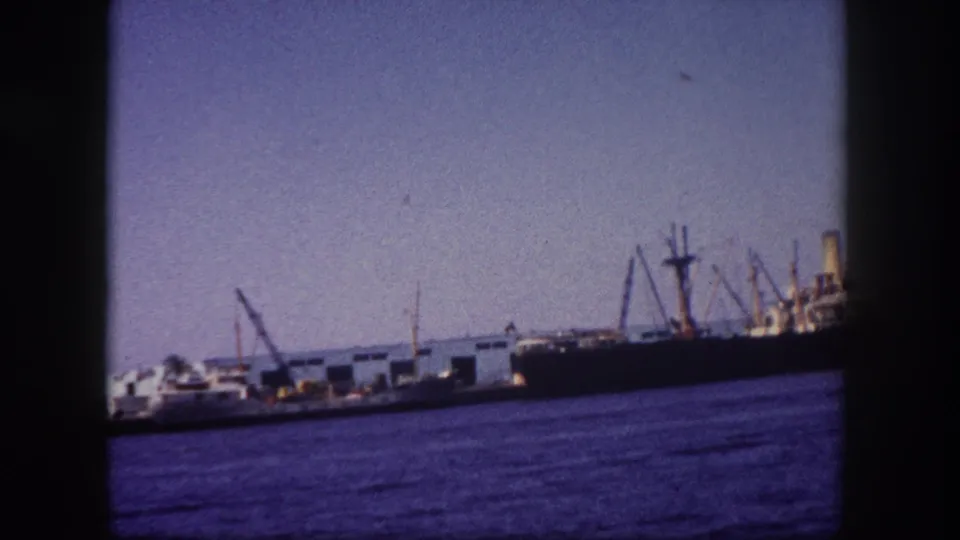 Cargo ship moored at Tampa port with loading derrick booms, circa 1960, blue-toned water in foreground, 8mm archival footage.
