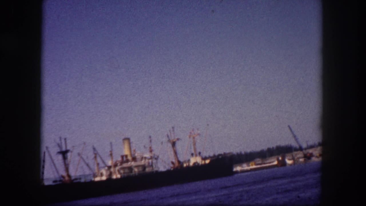 Wide harbor view with several cargo ships and boom-derrick cranes moored at a pier, 8mm film.
