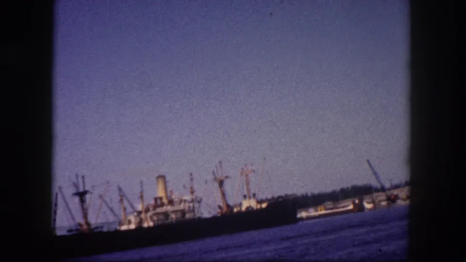 Wide 8mm harbor shot of multiple cargo ships with boom-derrick cranes at a pier, Tampa, Florida, circa 1960, with color-shifted blue water and hazy sky.