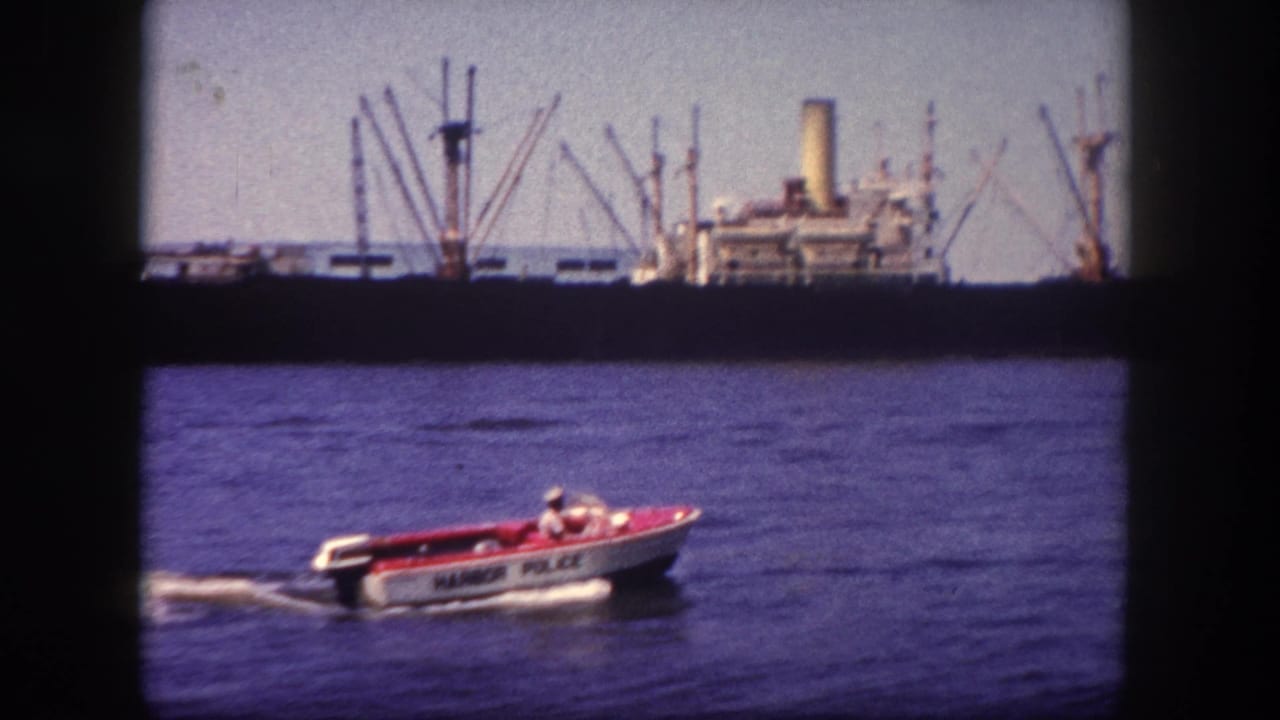 Red and white Harbor Police speedboat crossing Tampa harbor water, circa 1960, with large cargo ship and boom-derrick cranes in the background.