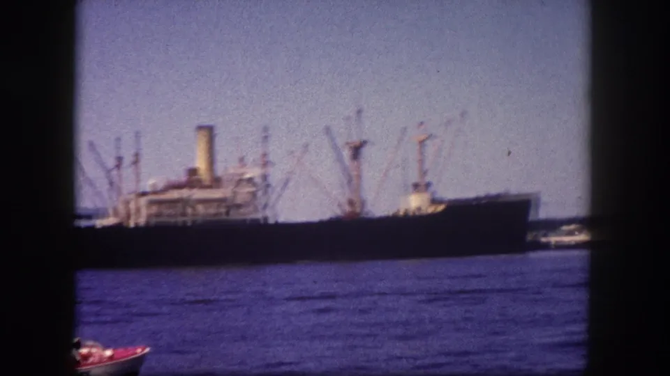 Large cargo ship with multiple boom-derrick cranes moored at Tampa harbor pier, circa 1960; a small red boat appears at the lower frame edge.
