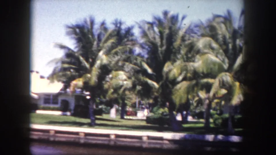 Closing shot of a mid-century waterfront property with red tile roof through coconut palms; a small outbuilding or structure appears at right of frame, Tampa area, circa 1960.