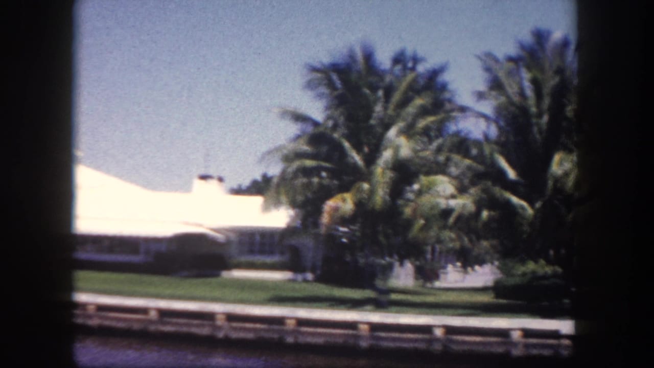 View from a boat of a white low-roofed home behind large palm trees and a green lawn, with a wooden waterfront seawall and blue canal.