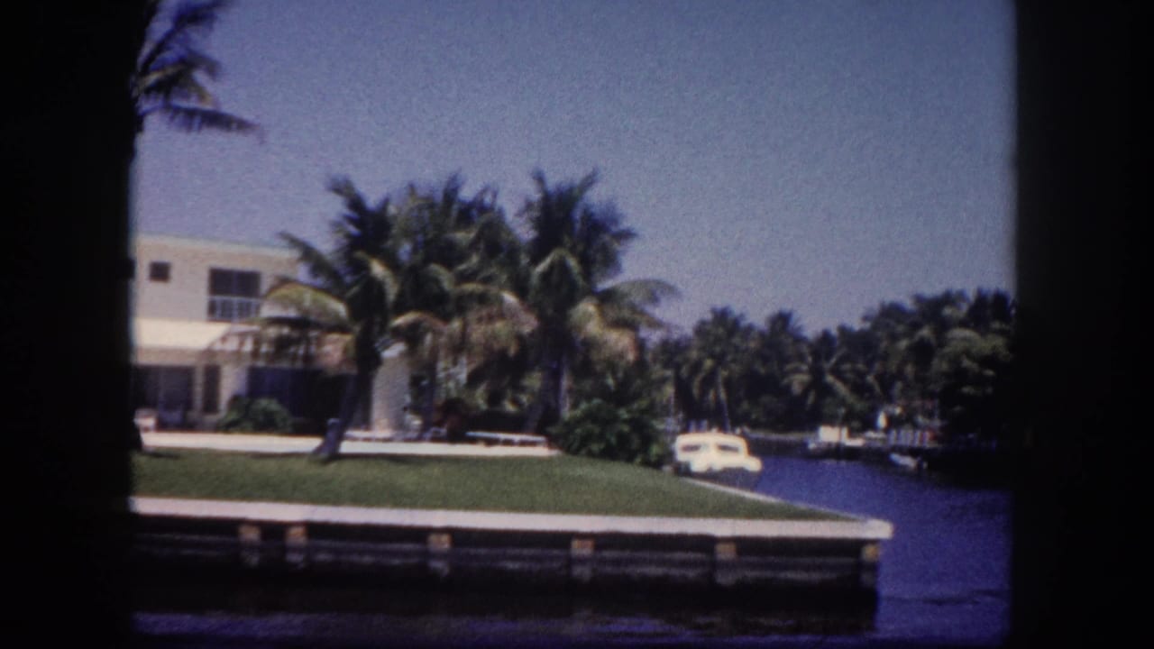 8mm film frame circa 1960 of a canal-front two-story Florida home with flat roof, palm trees, a parked vehicle, and a wooden dock, identified as the Tampa area.