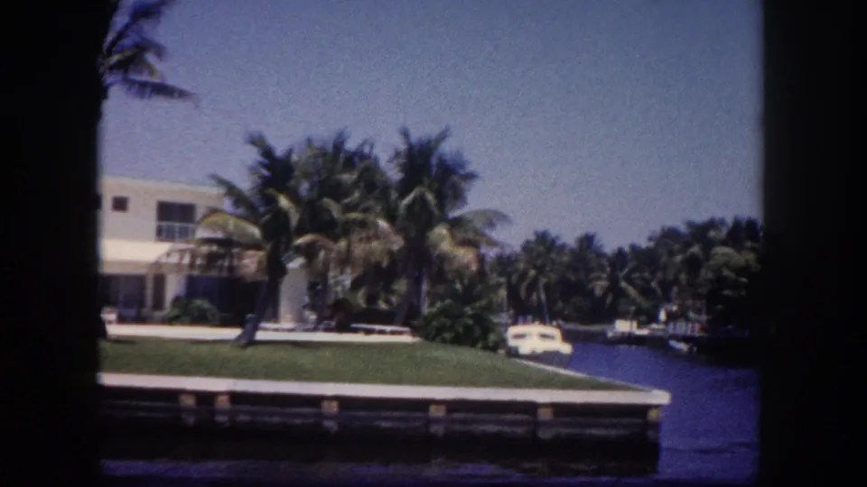 8mm film frame circa 1960 of a canal-front two-story Florida home with flat roof, palm trees, a parked vehicle, and a wooden dock, identified as the Tampa area.