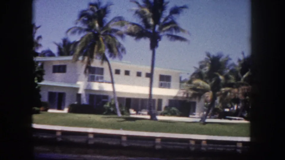8mm home-movie frame circa 1960 of a white mid-century modern canal-side house in the Tampa area, viewed from a passing boat, with tall palms and a wooden seawall.